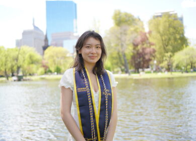 Woman wearing graduation stole standing in front of lake with skyscrapers in the backdrop.