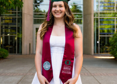 Woman standing with graduation cap and stole in front of University of Oregon logo.