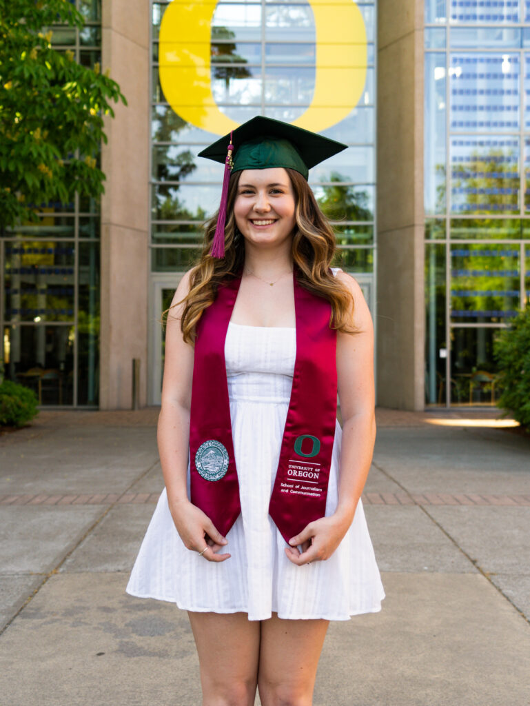 Woman standing with graduation cap and stole in front of University of Oregon logo.