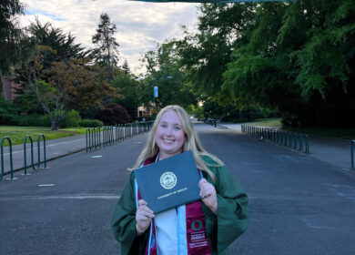 Intern Samantha Wettstein dressed in graduation robe, posing by trees and sunset.