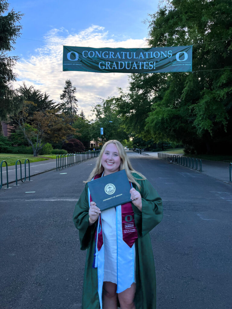 Intern Samantha Wettstein dressed in graduation robe, posing by trees and sunset.