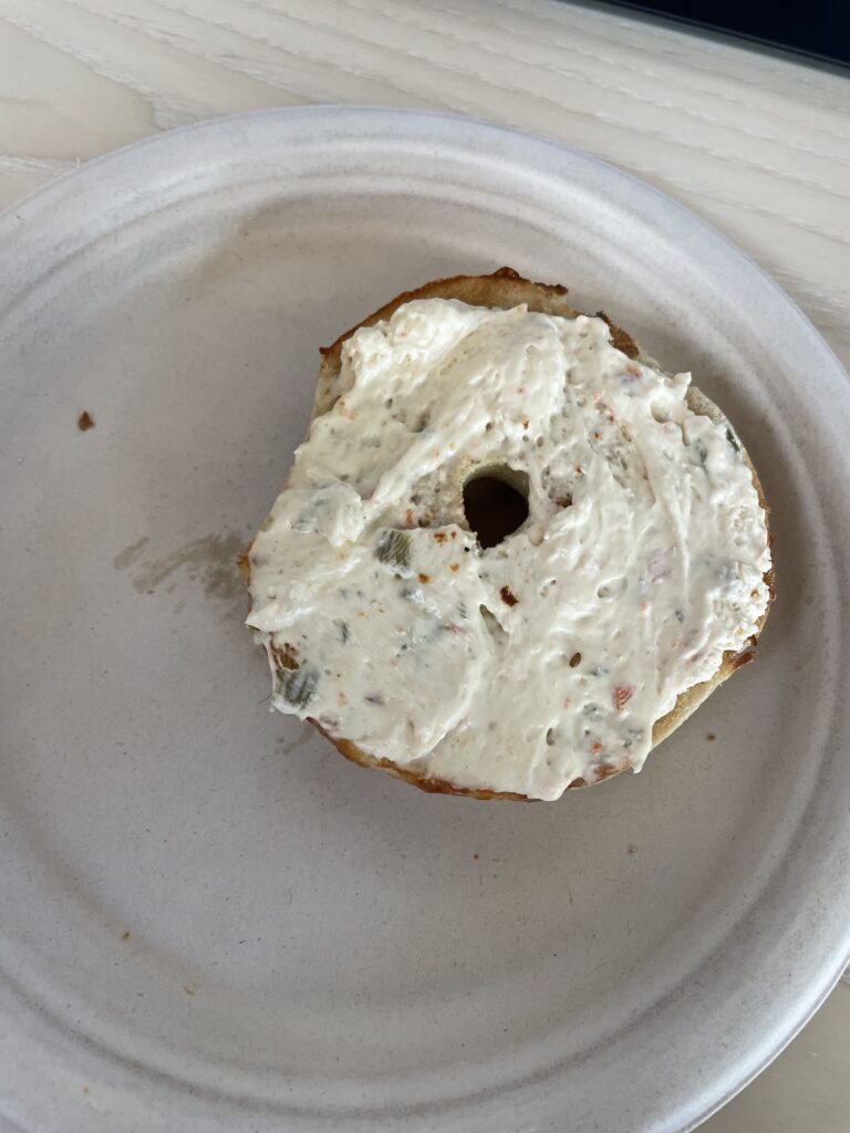 Half of bagel, covered in cream cheese, sitting on a styrofoam plate.