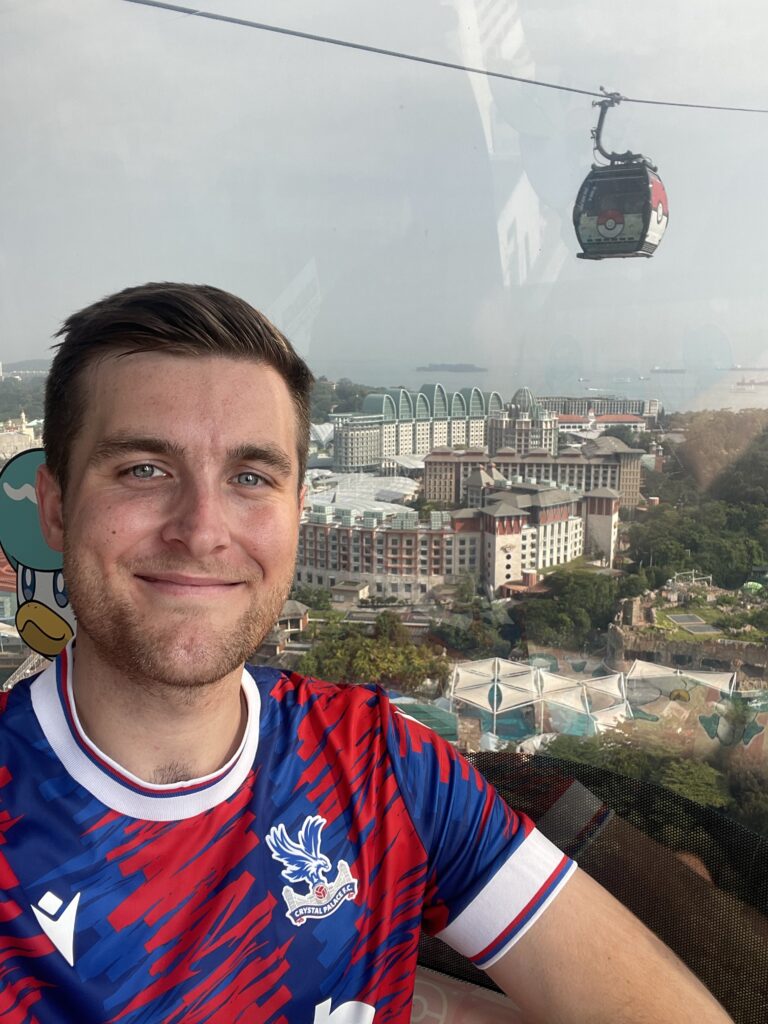 Man in bright blue and red shirt standing in front of cable car and scenic overlook.