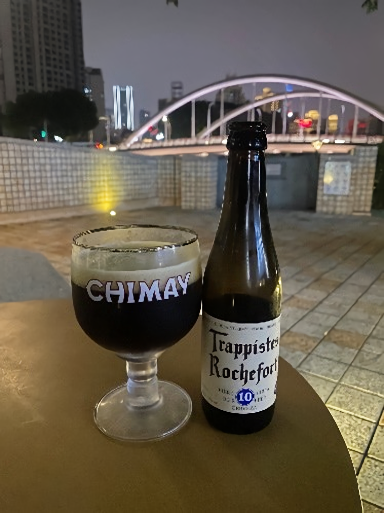 Glass full of dark beer sitting next to Belgian beer bottle on wood table outside.