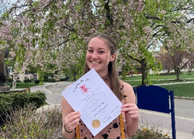 Woman standing outdoors by trees and grass, holding a diploma.