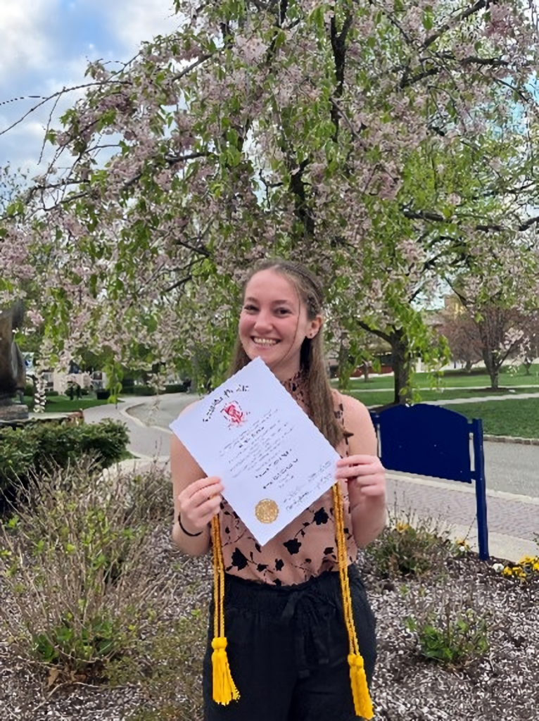 Woman standing outdoors by trees and grass, holding a diploma.