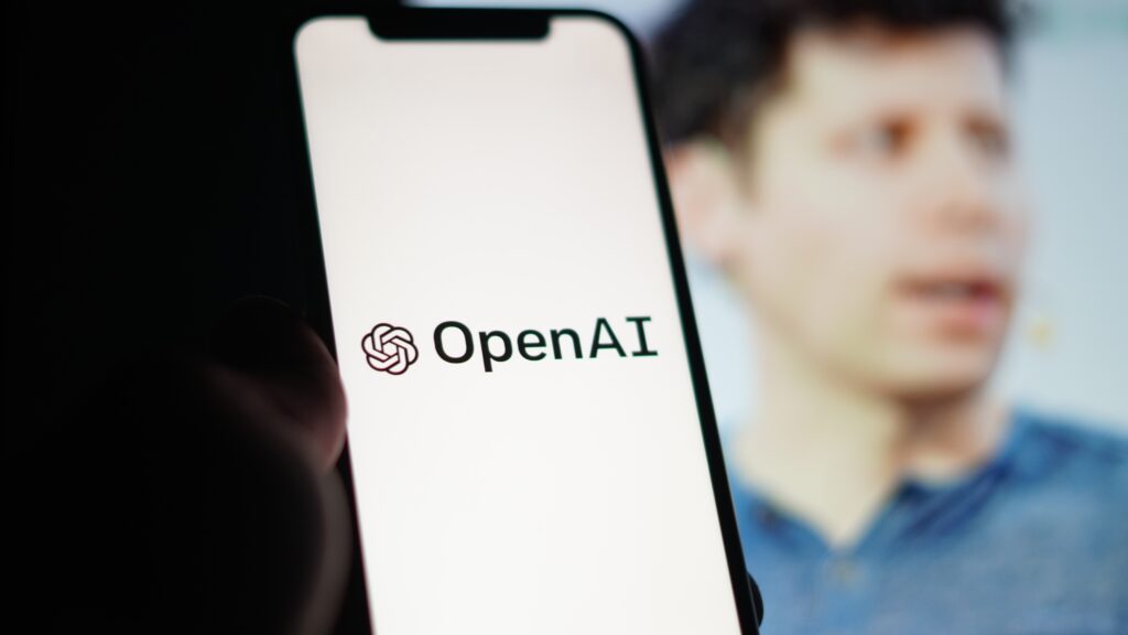 Man wearing blue shirt holding a mobile phone toggled to an OpenAI launch screen.