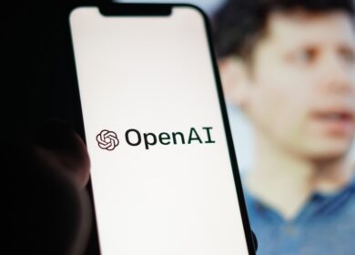 Man wearing blue shirt holding a mobile phone toggled to an OpenAI launch screen.