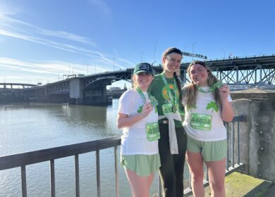 Three women wearing race medals and athletic clothes, standing in front of a river.